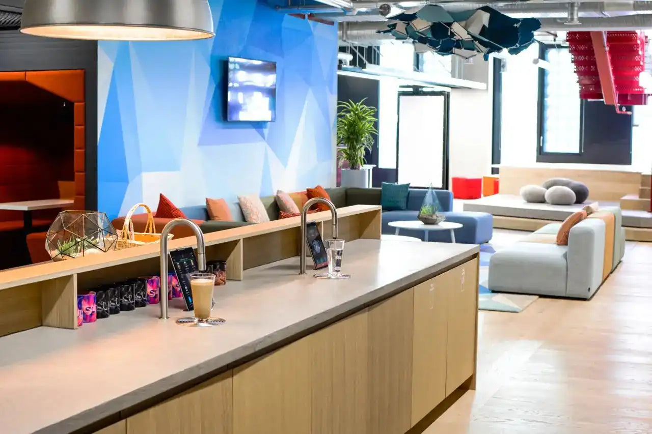 A contemporary office lounge with varied seating, a long counter with drink dispensers, and a geometric blue accent wall.