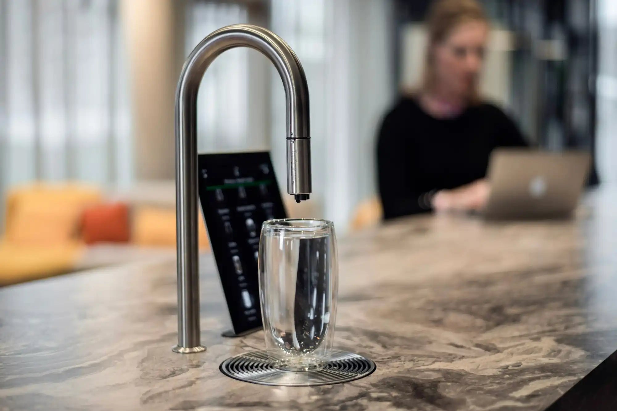 A modern water dispenser with a sleek, U-shaped stainless steel faucet and a digital control panel, filling a clear glass.